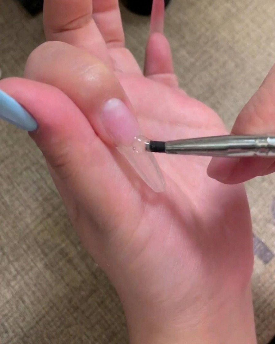 Ein Pinsel trägt eine Schicht hautfarbenes Gel auf einen vorbereiteten Nagel auf.