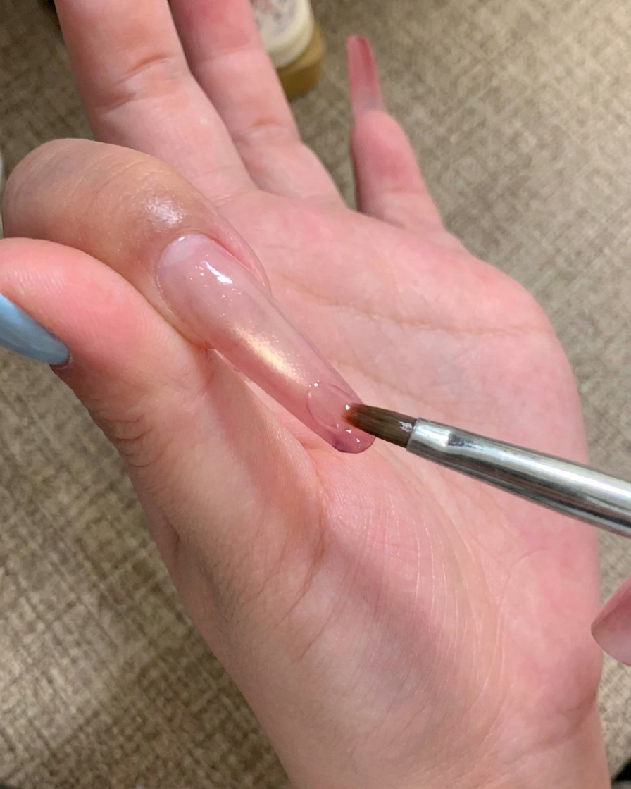 Ein Nail-Art-Pinsel trägt einen Tropfen klares Klebegel auf die Spitze eines langen Nagels auf.