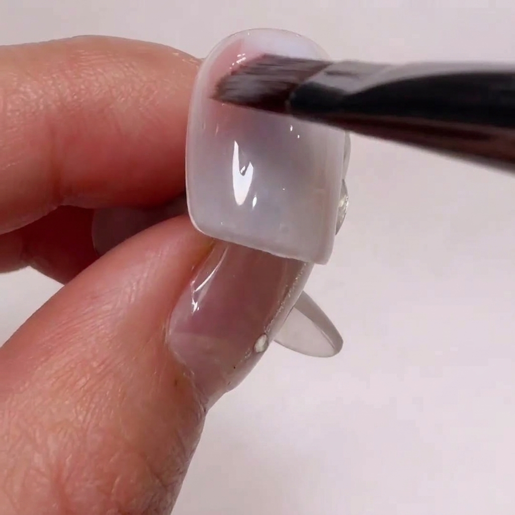 Ein Nagelkunst-Pinsel trägt ein sanftes Nude-Rosa-Gel auf die Ecken eines Nagel-Tips auf.