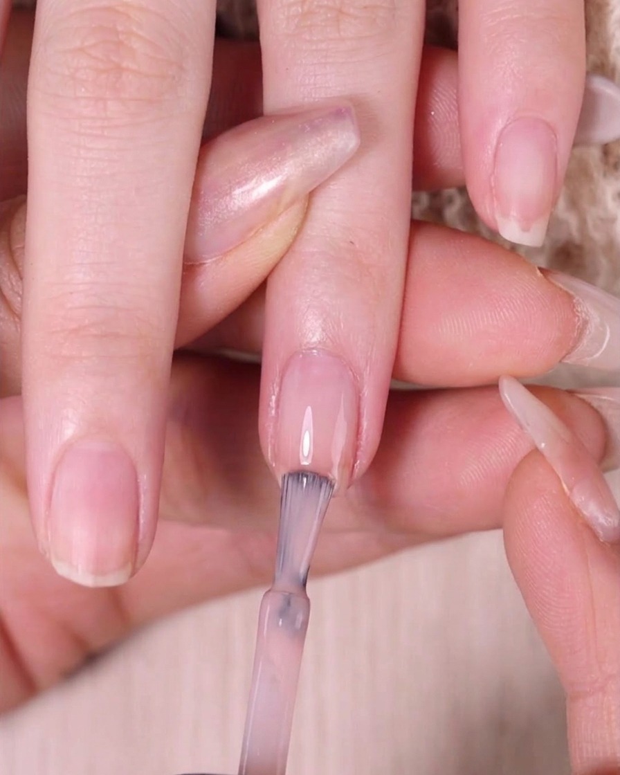 Ein Nagellackpinsel trägt transparenten nudefarbenen Lack nur auf das obere Drittel des Nagels auf.
