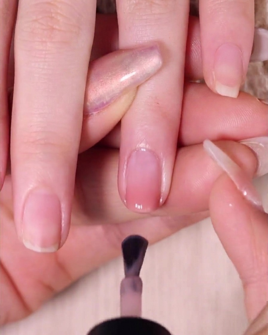 Ein Pinsel trägt eine glatte Schicht transparenten Nude-Pink-Lacks über den Ombré-Nagel auf, lässt jedoch einen kleinen Abstand zur Nagelhaut.