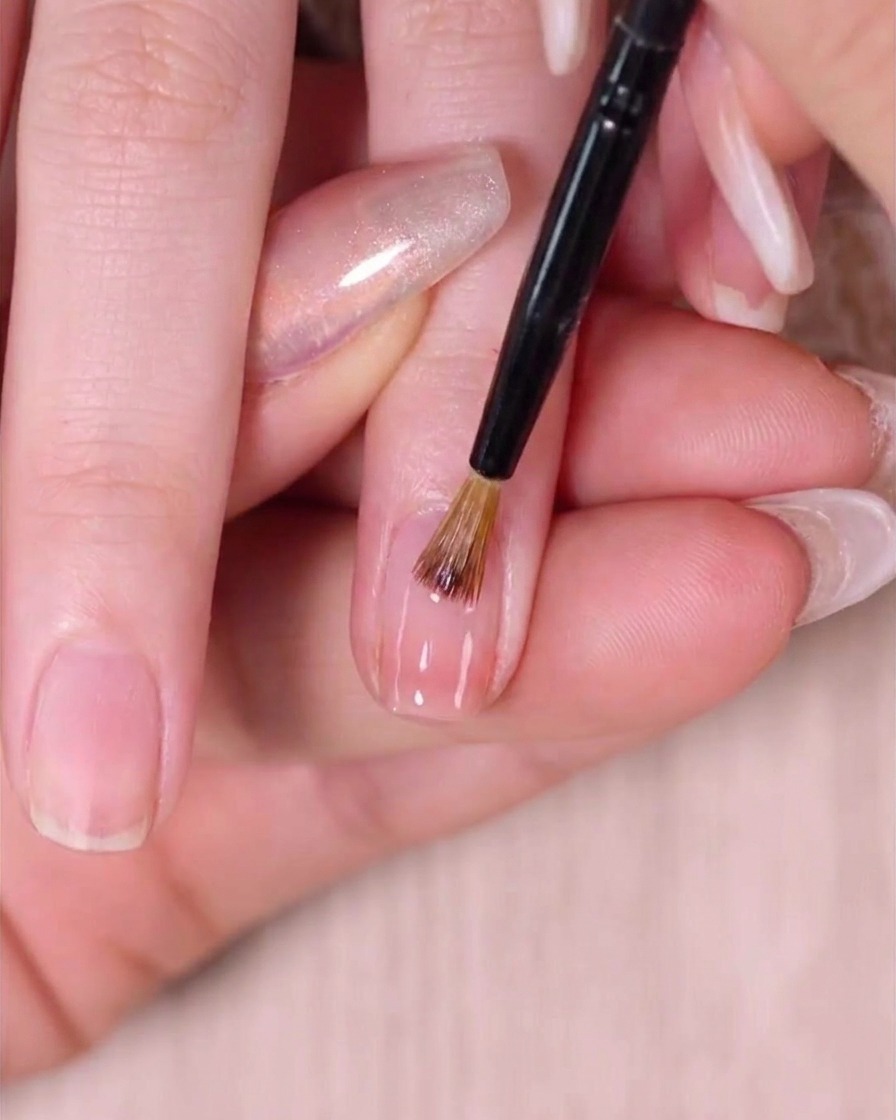 Ein feiner Nail-Art-Pinsel verblendet sanft feuchten rosa Lack nahe der Nagelhaut für einen nahtlosen Übergang.