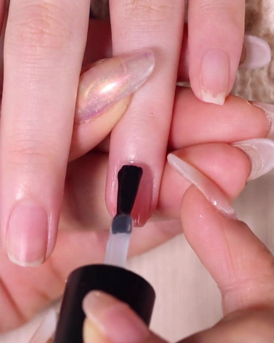 Ein Pinsel trägt eine dicke, glänzende Schicht klaren Überlacks auf einen fertigen Nude-Pink-Ombré-Nagel auf.