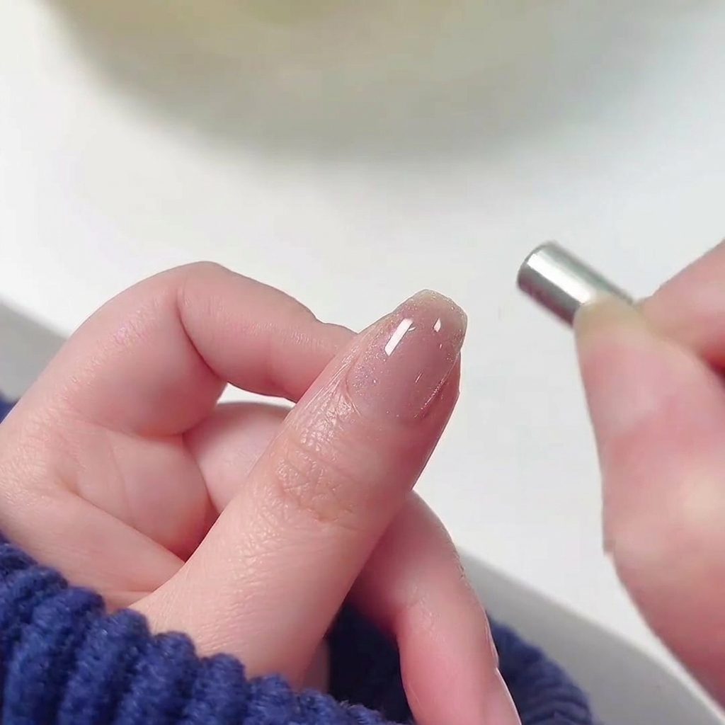 Ein kleiner zylindrischer Magnet, der in der Nähe der Nagelhaut eines mit feuchtem, schimmerndem rosa Cat-Eye-Gel lackierten Fingernagels gehalten wird.