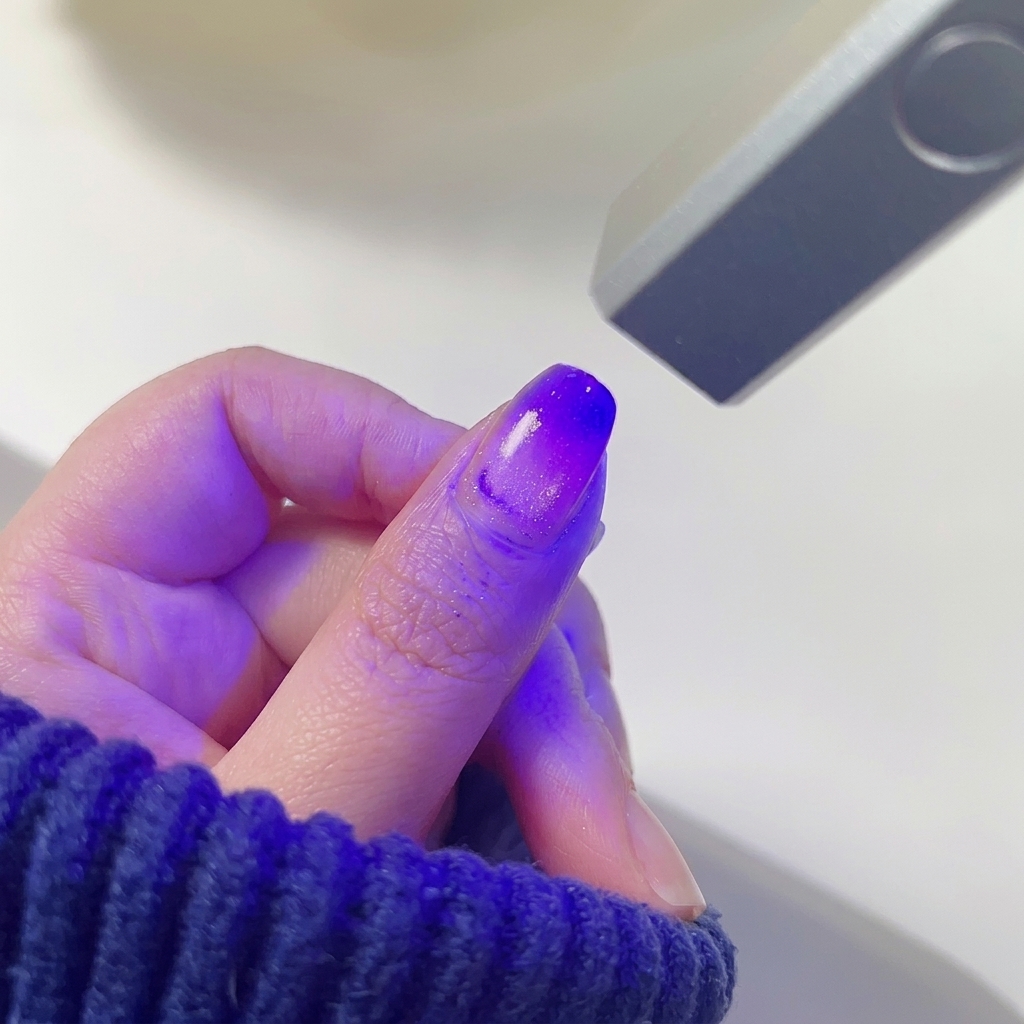 Ein Daumennagel mit einem verblendeten pink-lila Farbverlauf, der unter dem violetten Licht einer Mini-LED-Nagellampe aushärtet.