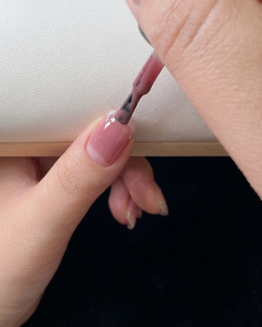 Nahaufnahme eines Pinsels, der eine zweite Schicht rosa Gel-Nagellack aufträgt und in Richtung Nagelbett verblendet.