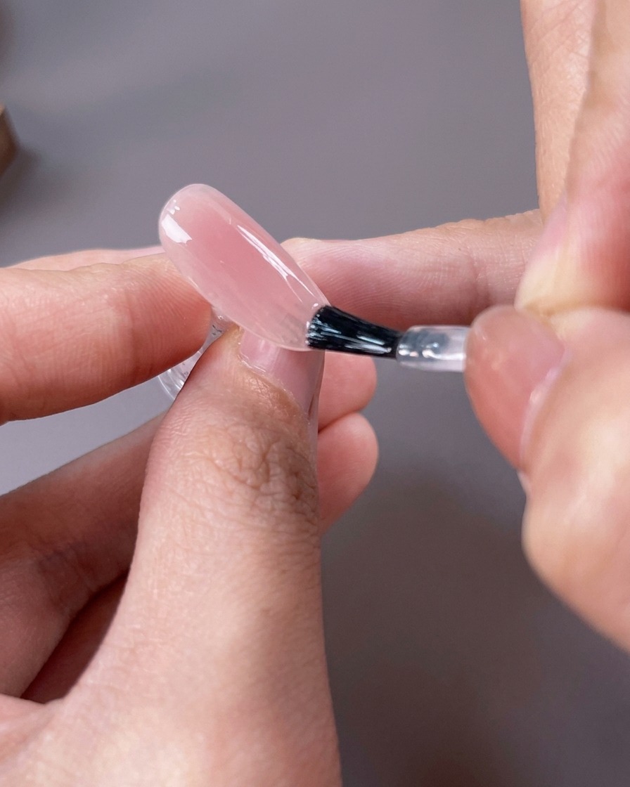 Ein Pinsel trägt einen transparenten, leicht rosafarbenen Nude-Gellack auf die gesamte Nageloberfläche auf.