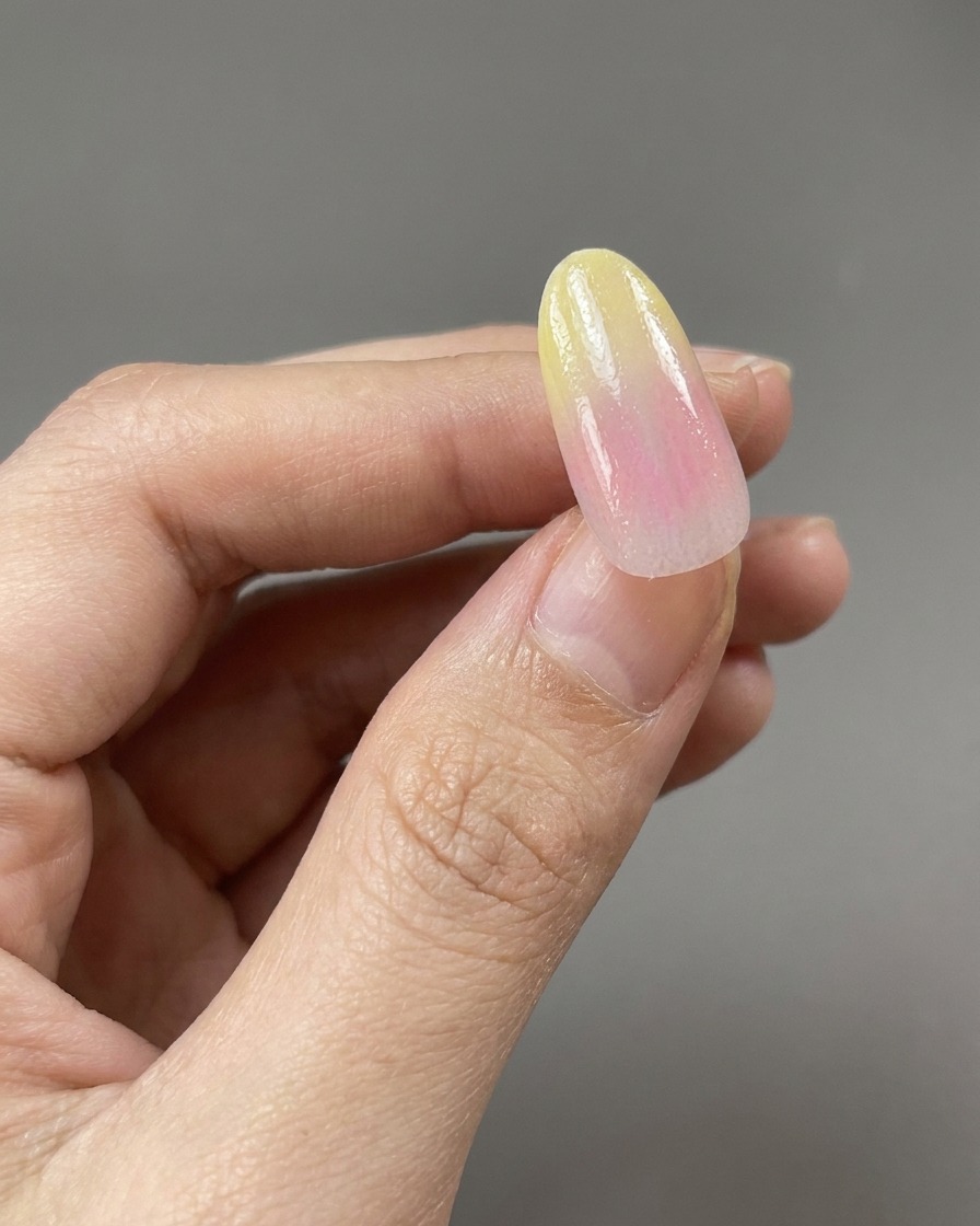 Ein Pinsel trägt einen klaren, glänzenden Base Coat über ein gelb-rosa Gradient-Nageldesign auf.