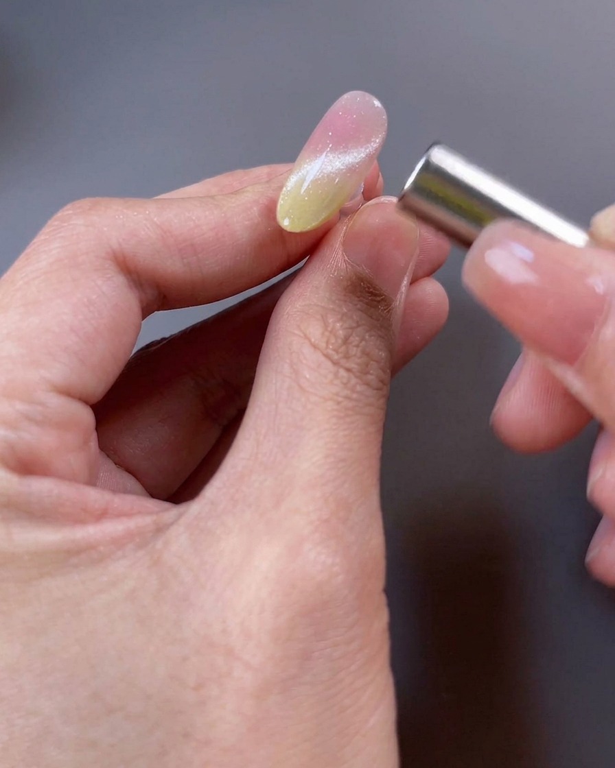 Nahaufnahme eines Nagels mit magnetischem Gellack. Ein Magnet wird über die Oberfläche gehalten, um die Schimmerpartikel zu einem breiten Lichtstrahl zu formen.