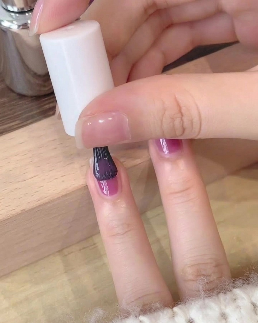 Nahaufnahme beim sorgfältigen Auftragen der zweiten Schicht tiefvioletten Gellacks auf einen Fingernagel.