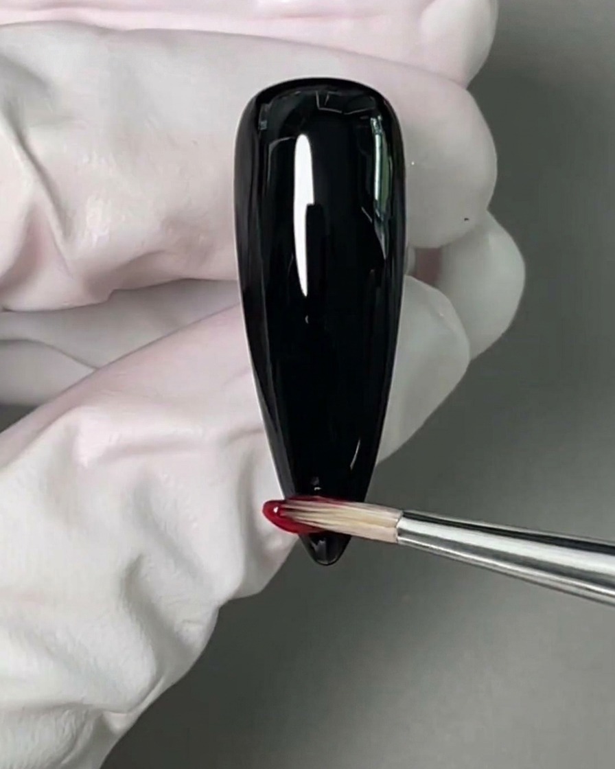 Ein kleiner Nailart-Pinsel trägt roten Gellack auf die Spitze eines schwarz lackierten Nagels auf.