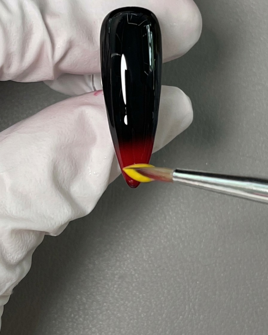 Ein Detailpinsel setzt einen Tropfen leuchtend gelben Gellack direkt über das nasse Rot an der Nagelspitze.