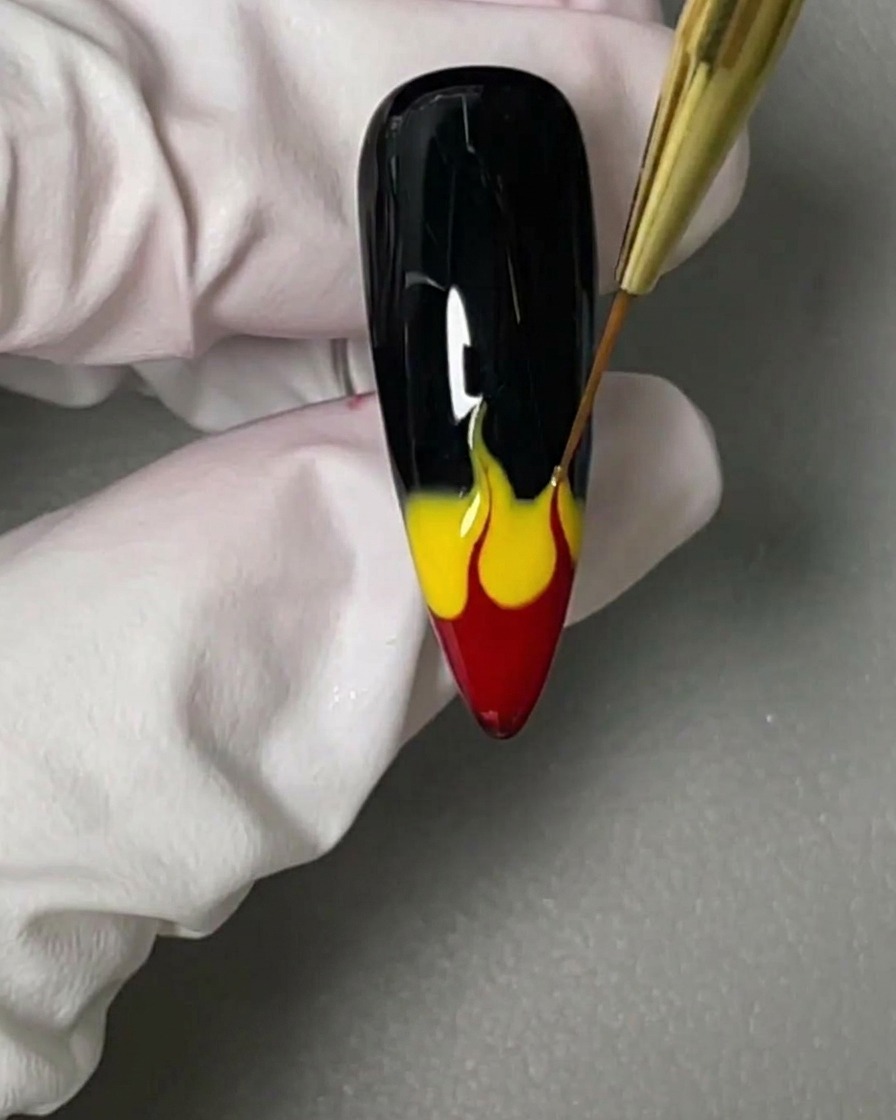 Ein feiner Liner-Pinsel zieht das nasse rote und gelbe Gel auf einem schwarzen Nagel nach oben zu Flammenformen.