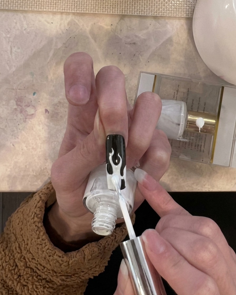 Ein Pinsel trägt eine dünne Schicht weißen Gellacks gezielt auf die Flammen-Designs an der Nagelspitze auf.