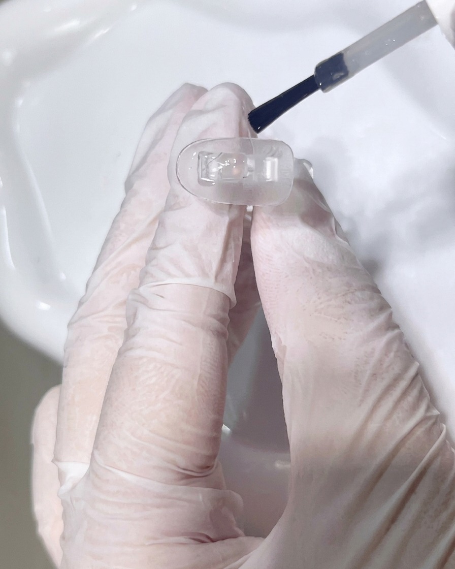 Nahaufnahme eines Pinsels, der einen klaren Base Coat auf einen transparenten Nagel-Tip aufträgt.