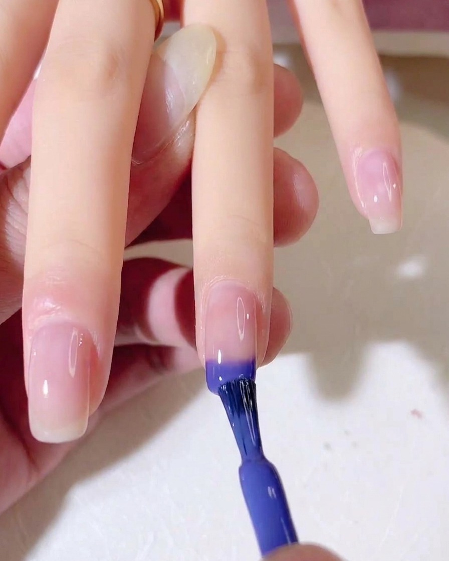 Ein Nagelpinsel trägt blaues und violettes Gel auf die Spitze eines natürlichen Fingernagels auf.