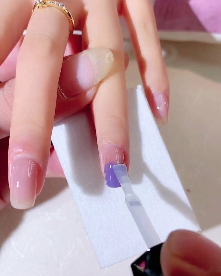 Ein Pinsel mit klarem Gel-Base-Coat verblendet die blauen und violetten Farben vertikal auf dem Nagel.