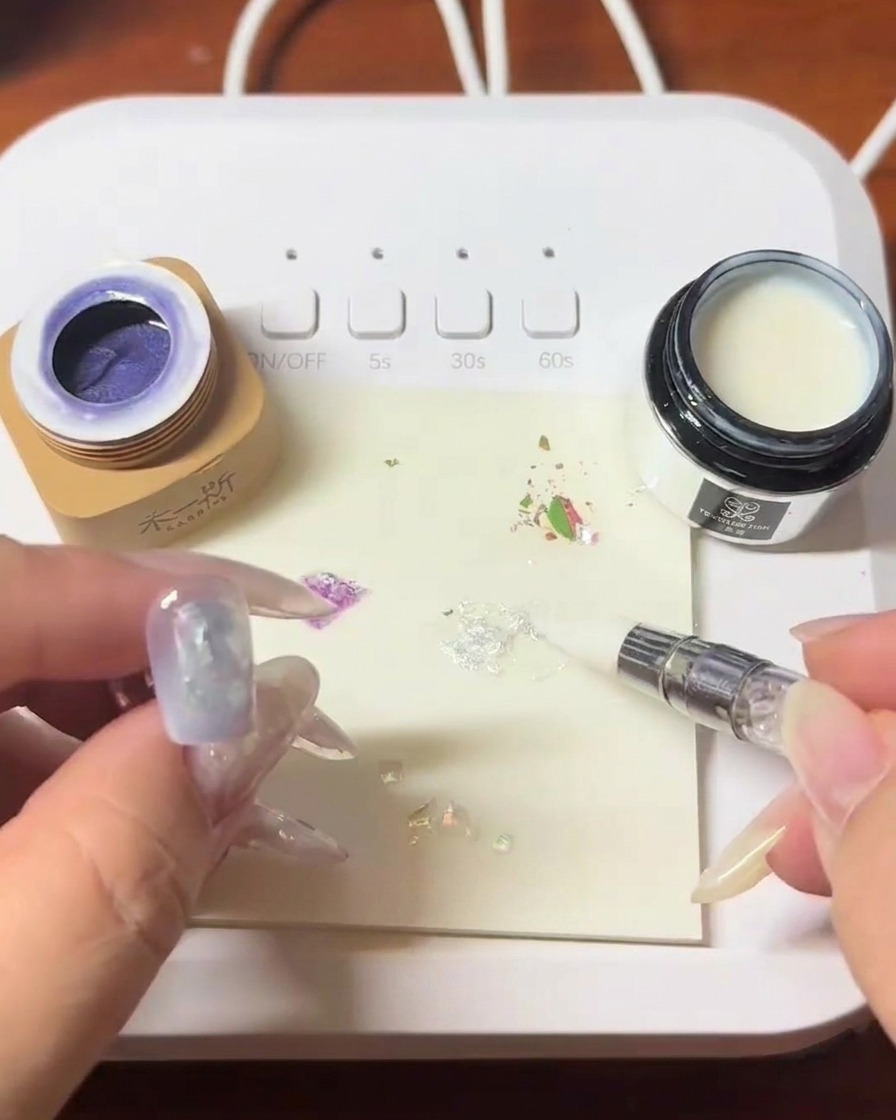 Ein Pinsel trägt klares Blooming-Gel über den mit Muscheln verzierten Nagel auf.