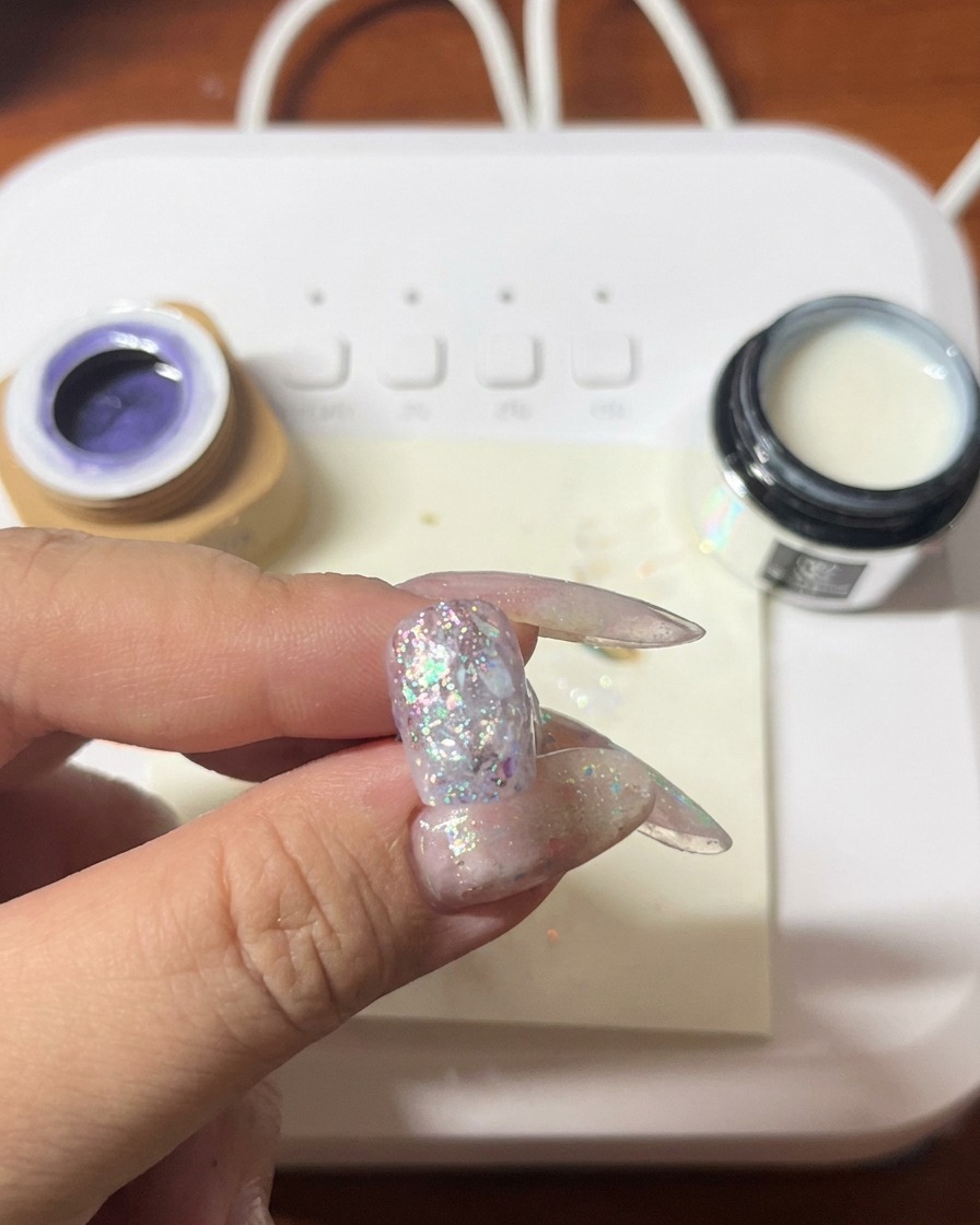 Ein Pinsel trägt violetten Gellack über einen bereits mit Glitzer und Muscheln verzierten Nagel auf.