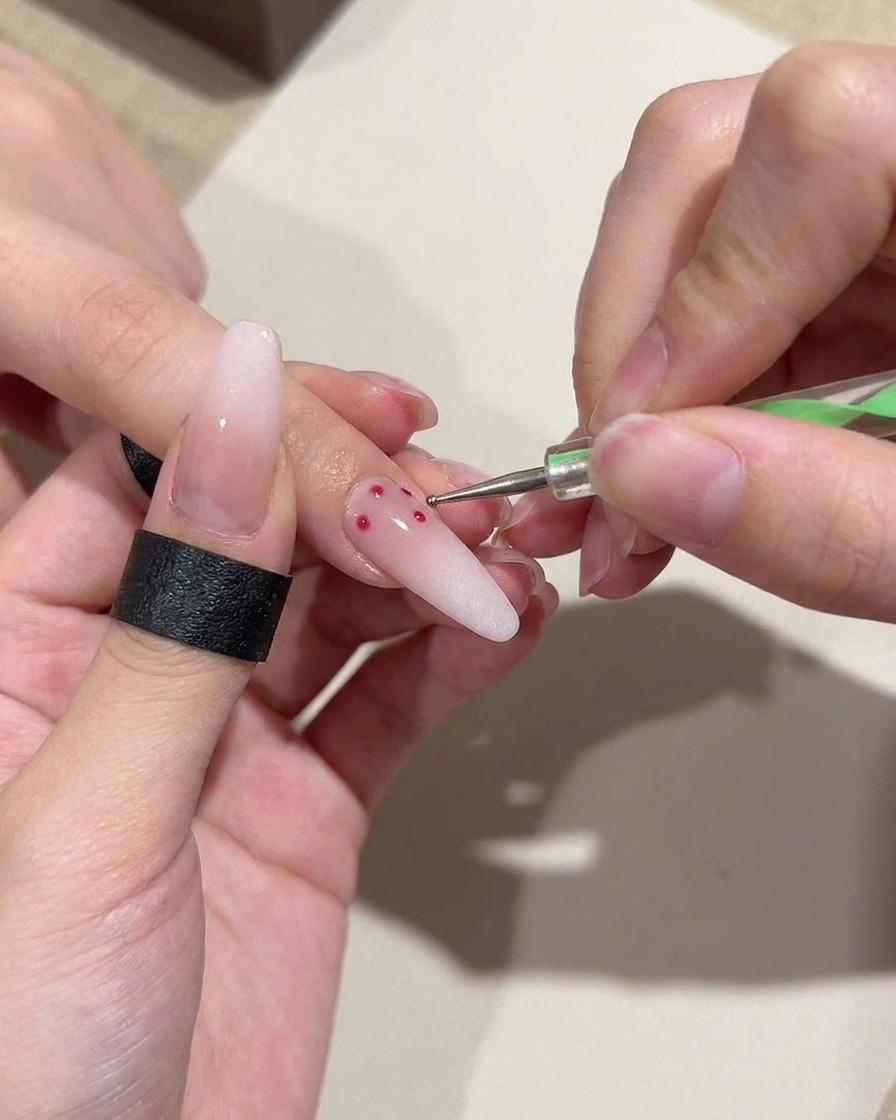 Nahaufnahme eines Dotting-Tools, das präzise, kleine rote Punkte auf einen zartrosa Gradient-Nagel setzt.