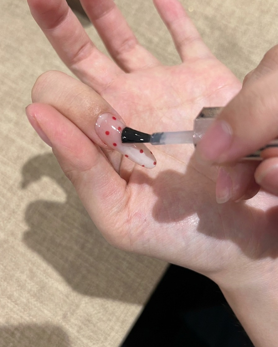 Nahaufnahme eines Nagelpinsels, der einen klaren, glänzenden Top Coat über einen mit roten Punkten verzierten Nagel aufträgt.