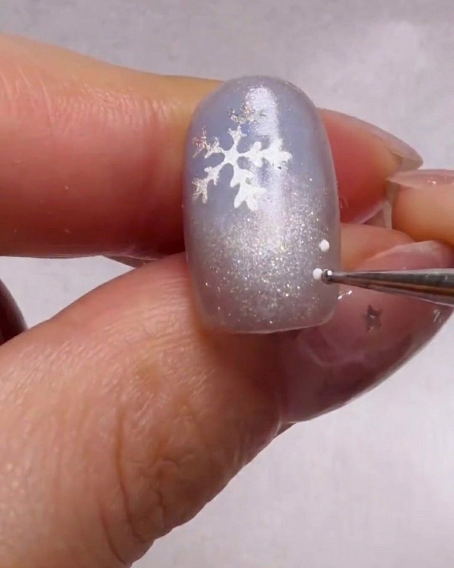 Nahaufnahme eines Dotting-Tools, das winzige weiße und hellblaue Punkte um einen silbernen Schneeflocken-Sticker auf einem schimmernden blauen Nagel setzt.