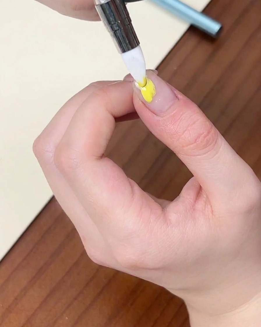 Ein feiner Liner-Pinsel trägt einen Punkt gelbes Gellack auf einen Naturnagel auf.