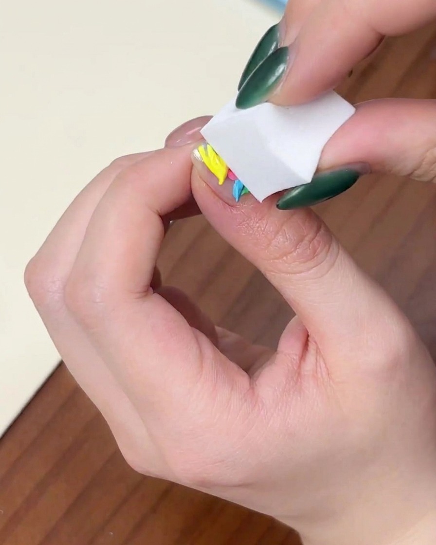 Nahaufnahme einer Hand, die ein weißes Kosmetikschwämmchen senkrecht auf einen Nagel mit bunten Gellack-Punkten drückt.