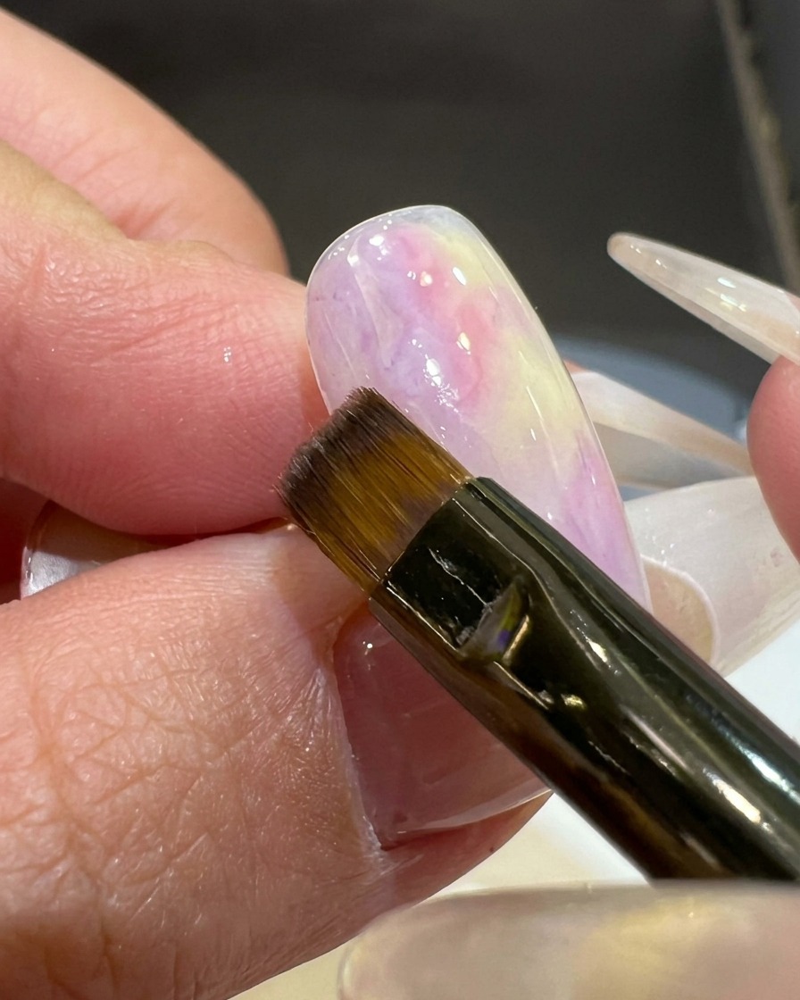 Ein flacher Pinsel trägt einen Hauch von rosa-violettem Gel auf die Ecke eines weiß grundierten Nageltips auf.
