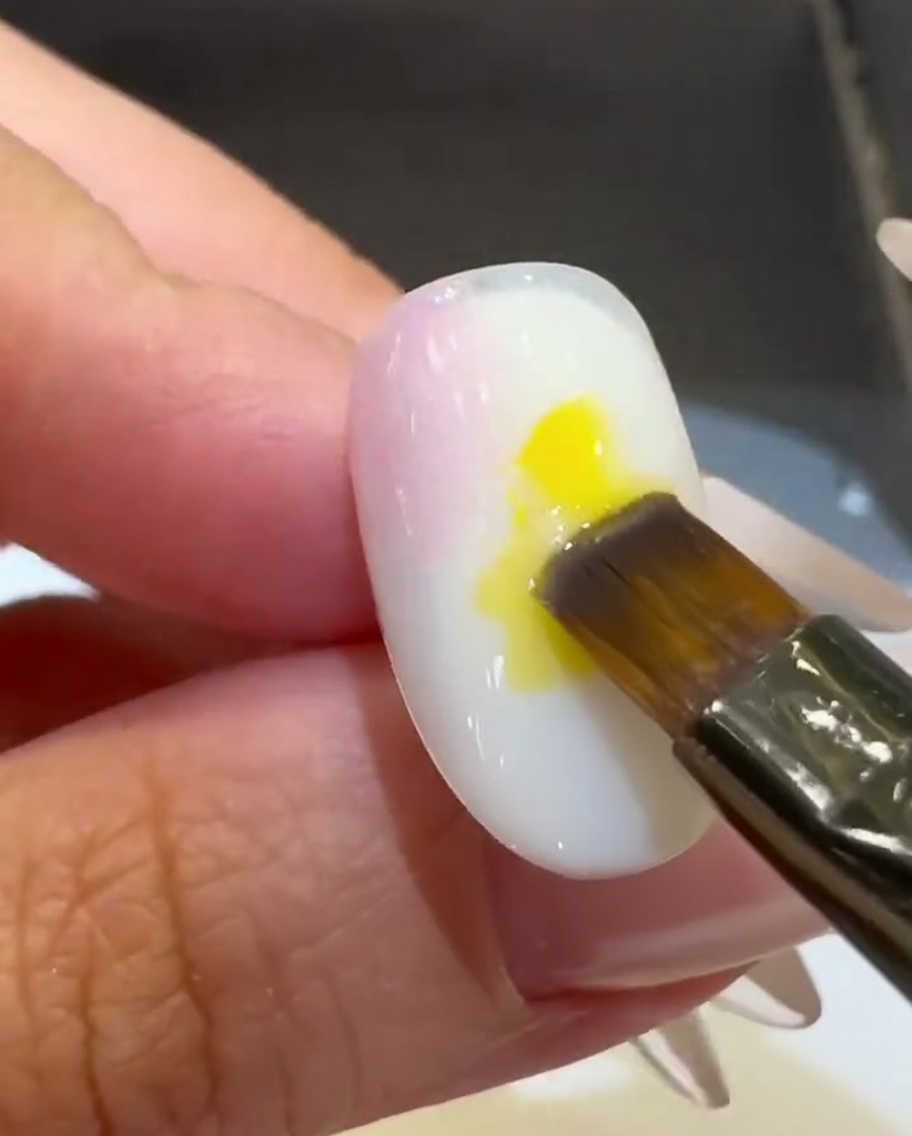 Ein Flachpinsel platziert einen Tropfen leuchtend gelben UV-Lack in der Mitte des Nagels.
