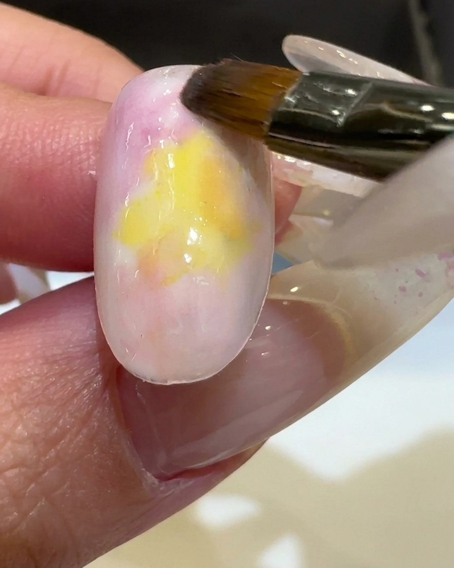 Ein Pinsel trägt leuchtend gelben UV-Lack in der Mitte eines Nagels auf und verblendet ihn.