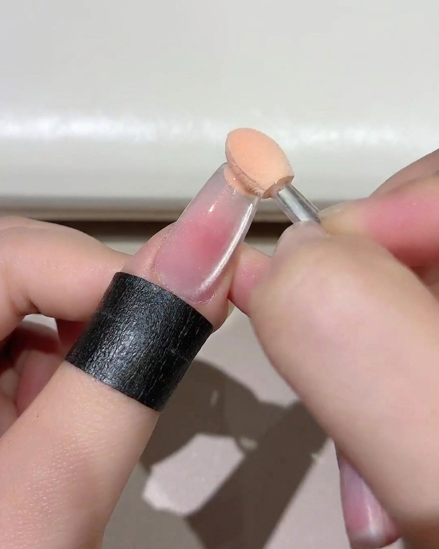 Nahaufnahme eines kleinen Schwämmchens, das pinkfarbenes Gel auf die freie Nagelspitze eines klaren Nagels aufträgt.
