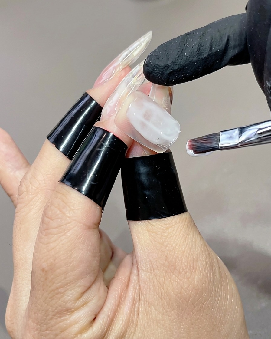 Eine Hand mit langen, klaren Nagelspitzen, auf deren Ringfinger ein flacher Pinsel einen milchig-weißen Gel-Base-Coat aufträgt.