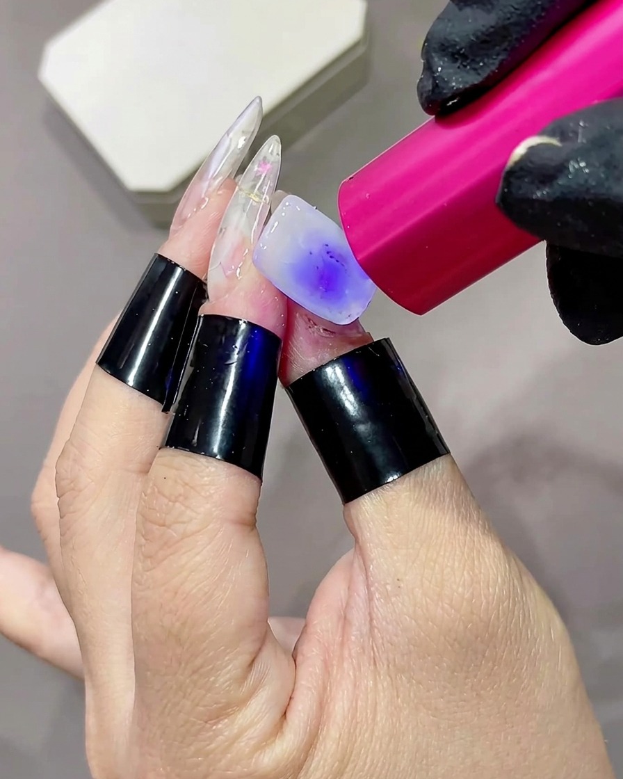 Nahaufnahme eines Fingers, dessen milchig-weißer Base Coat unter einer kleinen rosafarbenen UV- oder LED-Handlampe ausgehärtet wird.