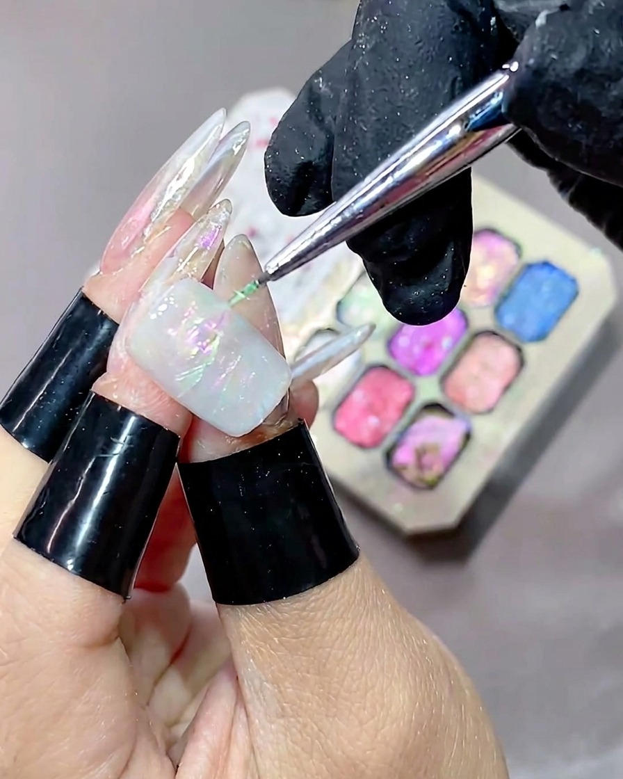 Ein feiner Liner-Pinsel trägt grünes Strukturgel über den Nagel auf, im unscharfen Hintergrund ist eine bunte Gelpalette sichtbar.