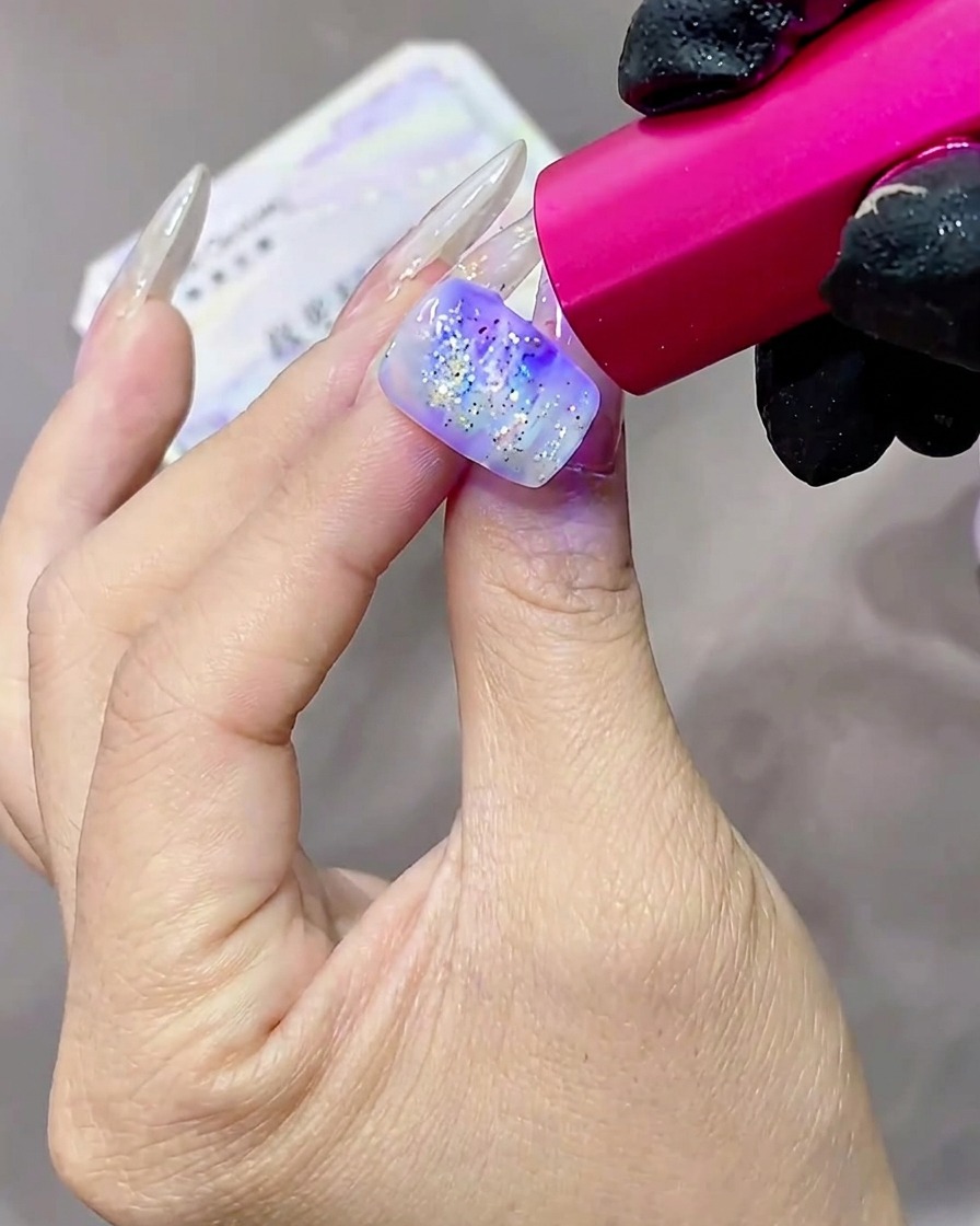 Eine Hand mit glitzernden, strukturierten Gelnägeln wird unter einer rosa getönten UV-Lampe ausgehärtet.