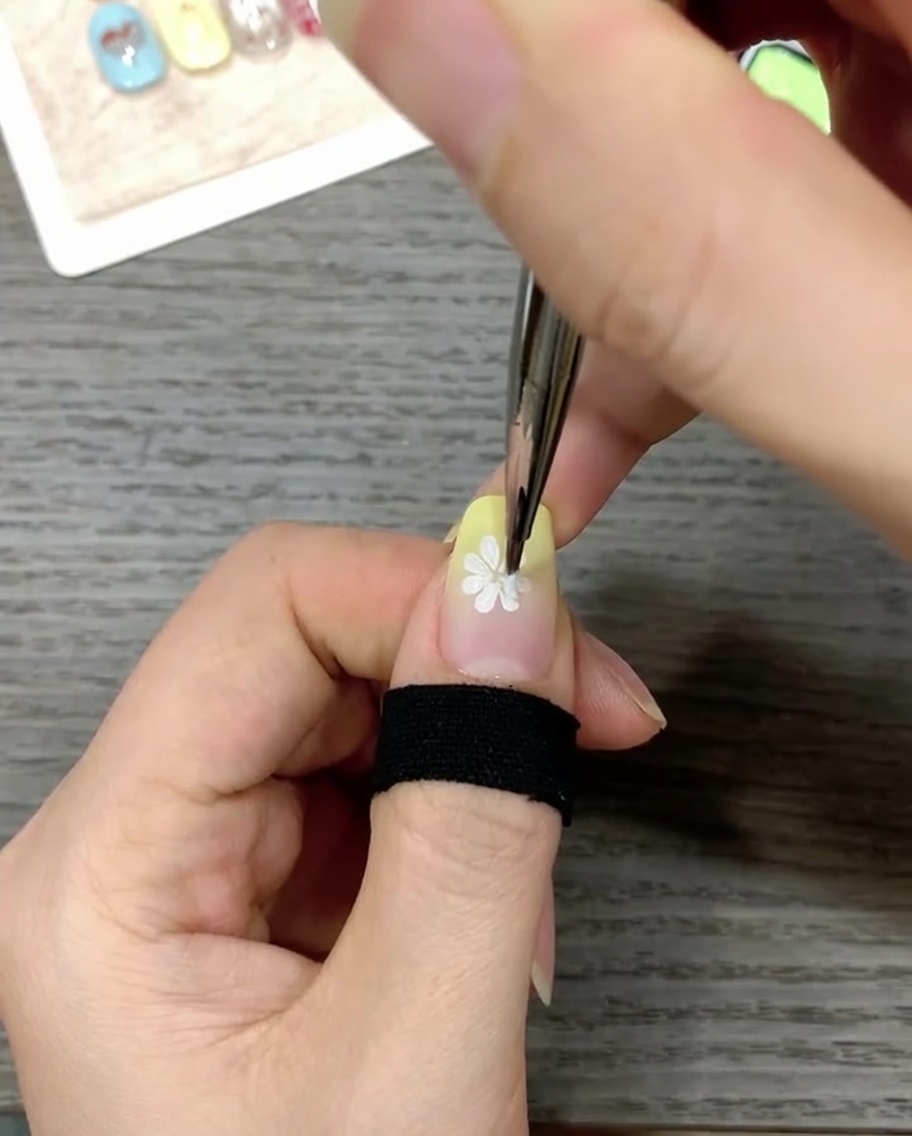 Nahaufnahme eines Nagels, bei dem mit einem feinen Pinsel ein kleiner, leuchtend gelber Punkt in die Mitte der weißen Blume gesetzt wird.