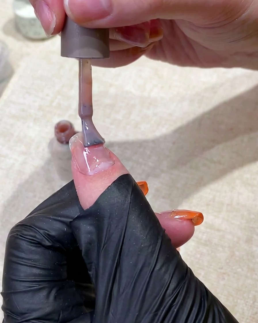 Eine Hand in einem schwarzen Handschuh trägt mit einem Pinsel einen durchscheinenden, nudefarbenen Base Coat auf den Fingernagel auf.