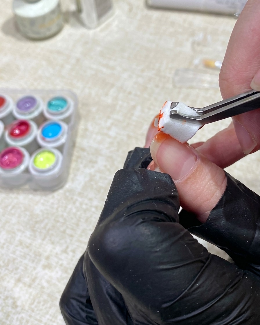 Eine behandschuhte Hand verwendet eine Pinzette, um orangefarbenen Gellack von einem kleinen Schwamm auf die Spitze eines Fingernagels zu tupfen.