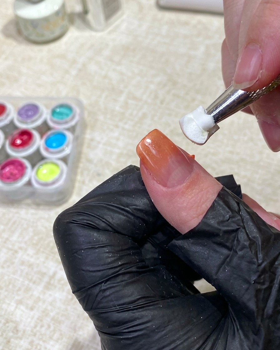 Ein Werkzeug hält ein sauberes weißes Schwämmchen und klopft sanft auf die Mitte eines Fingernagels, um orangefarbenen Gellack in eine Nude-Basis einzuarbeiten.
