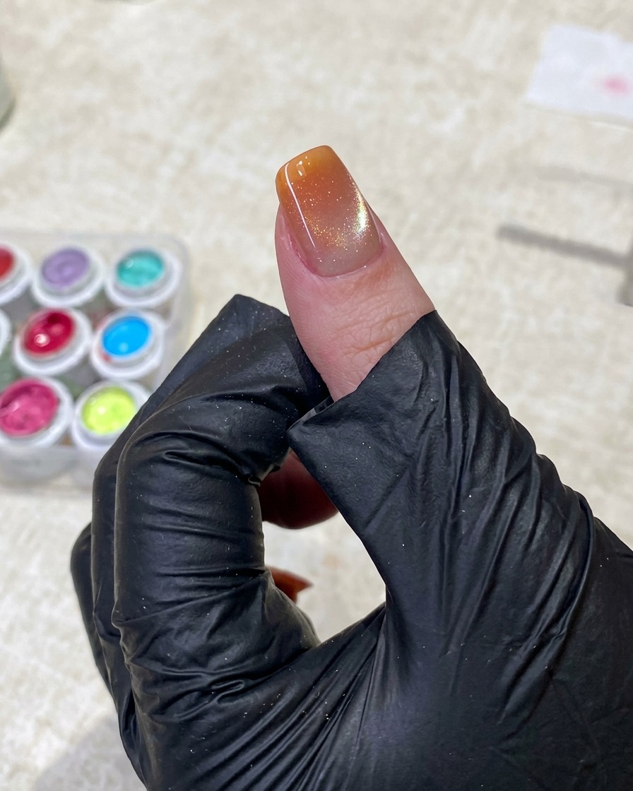 Eine Hand mit schwarzem Handschuh hält mit einer Pinzette einen kleinen Schwammapplikator und tupft sanft leuchtend orangefarbenen Gellack auf die Spitze eines manikürten Nagels.