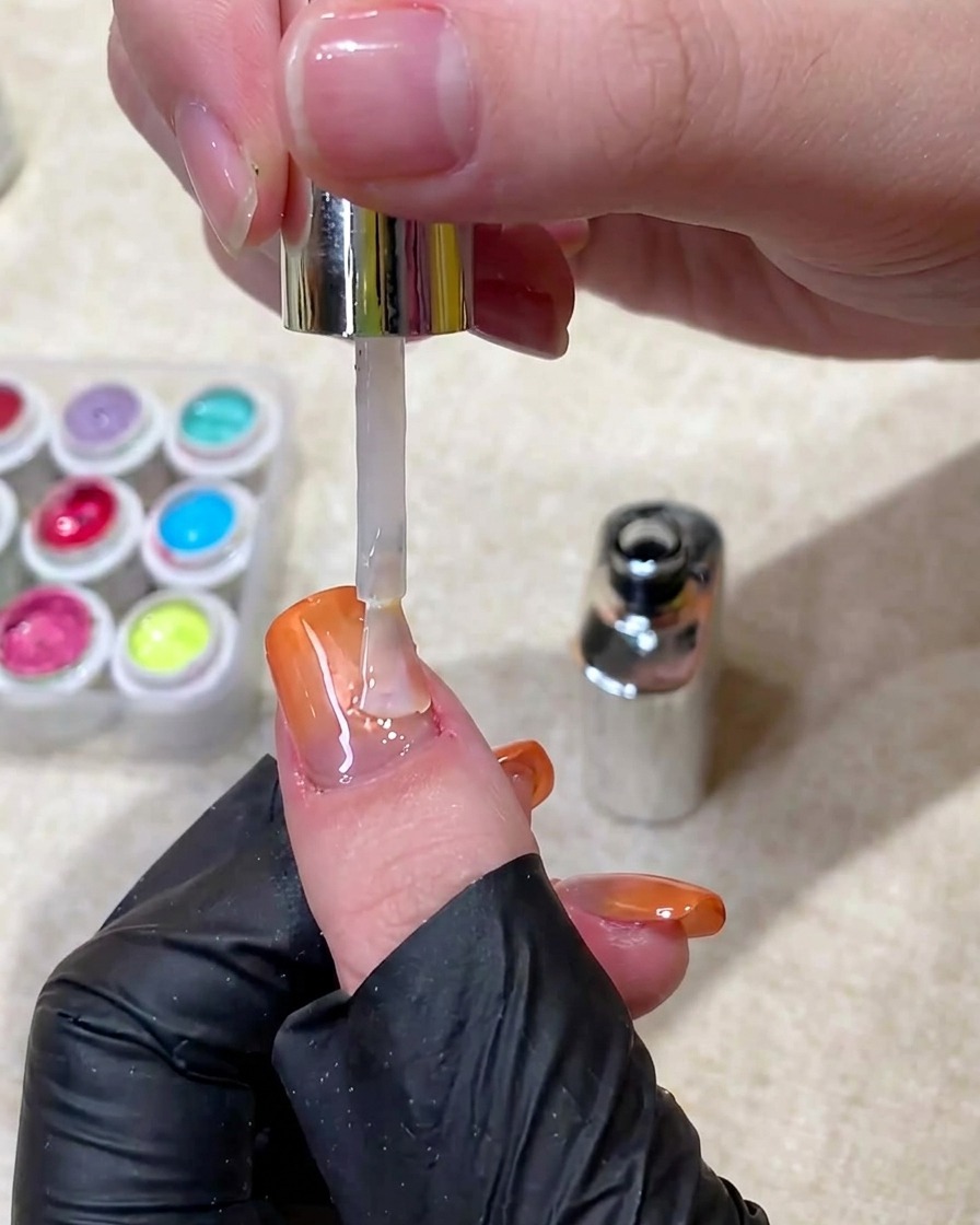Ein Pinsel trägt einen klaren, glänzenden Top Coat über einen fertigen, orangefarbenen Ombré-Cat-Eye-Nagel auf.
