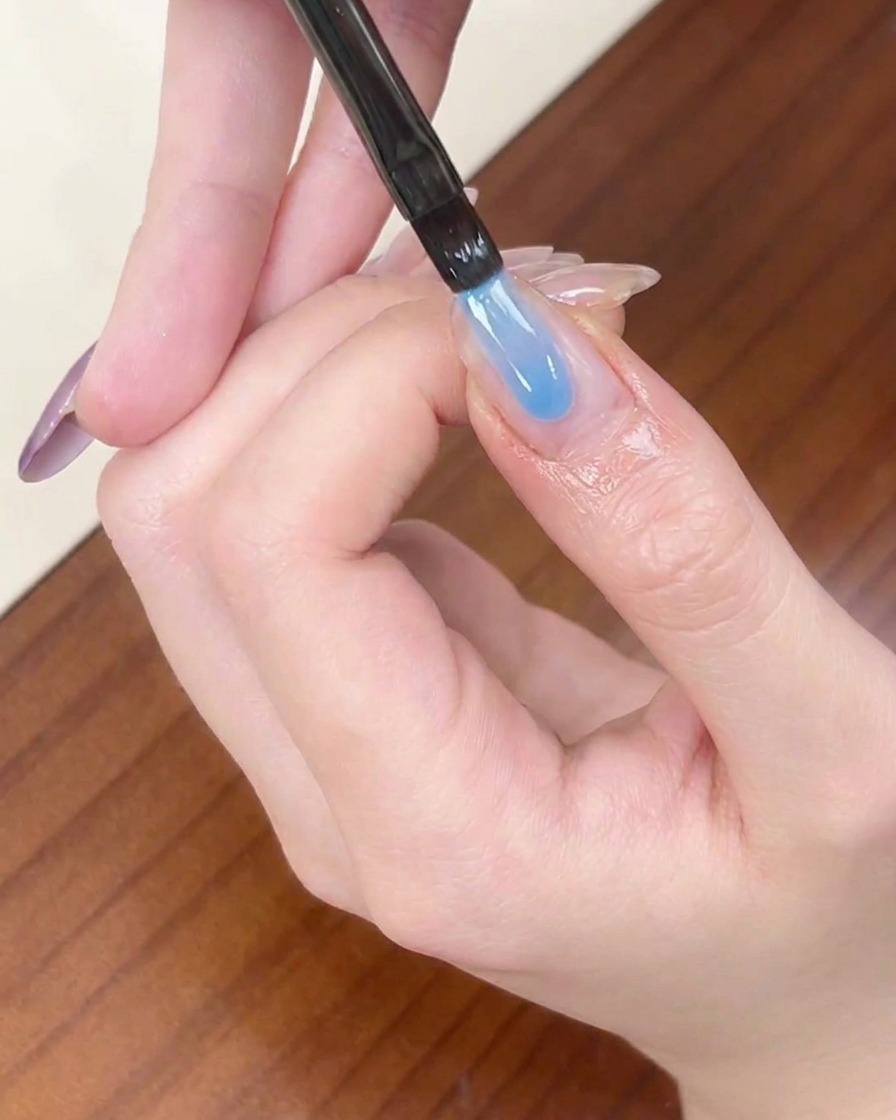 Ein Liner-Pinsel trägt einen Streifen des transparenten blauen Farbgels auf die Spitze eines mandelförmigen Fingernagels auf.