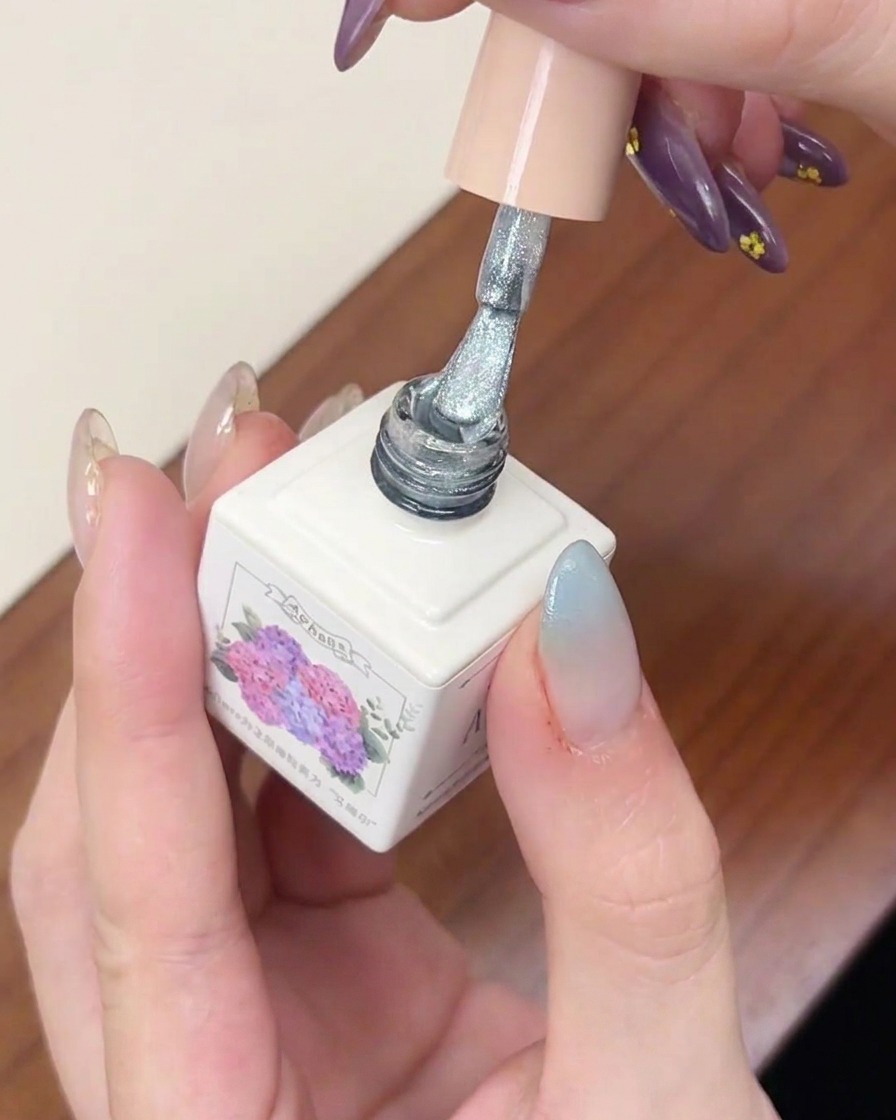 Ein Pinsel trägt eine Schicht silbern schimmernden Cat-Eye-Lack über einem Fingernagel mit blauem Farbverlauf auf.