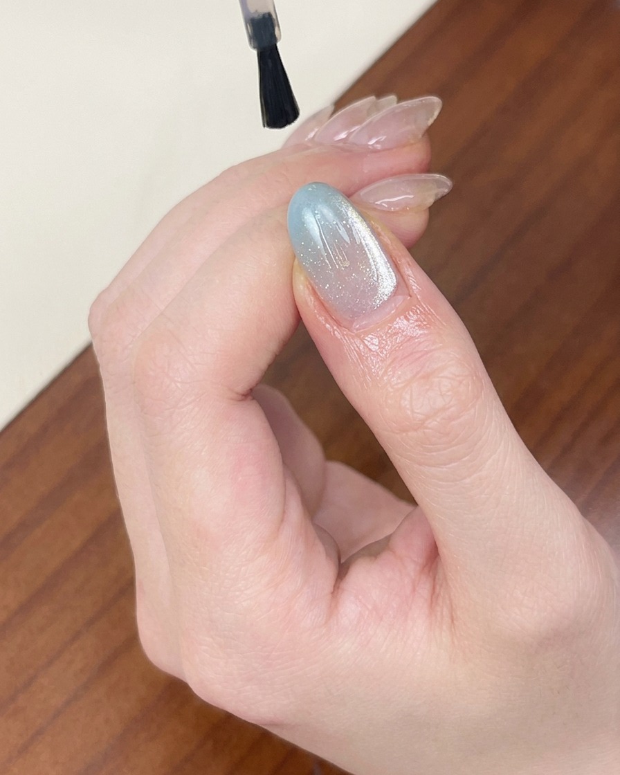 Ein Pinsel trägt einen klaren, glänzenden Top Coat über einem hellblauen, schimmernden Cat-Eye-Nagel auf.