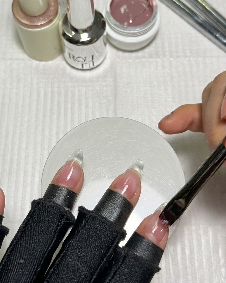 Ein Pinsel trägt eine glatte Schicht rosafarbenes Nude-Gel auf einen klaren, mandelförmigen Kunstnagel auf.