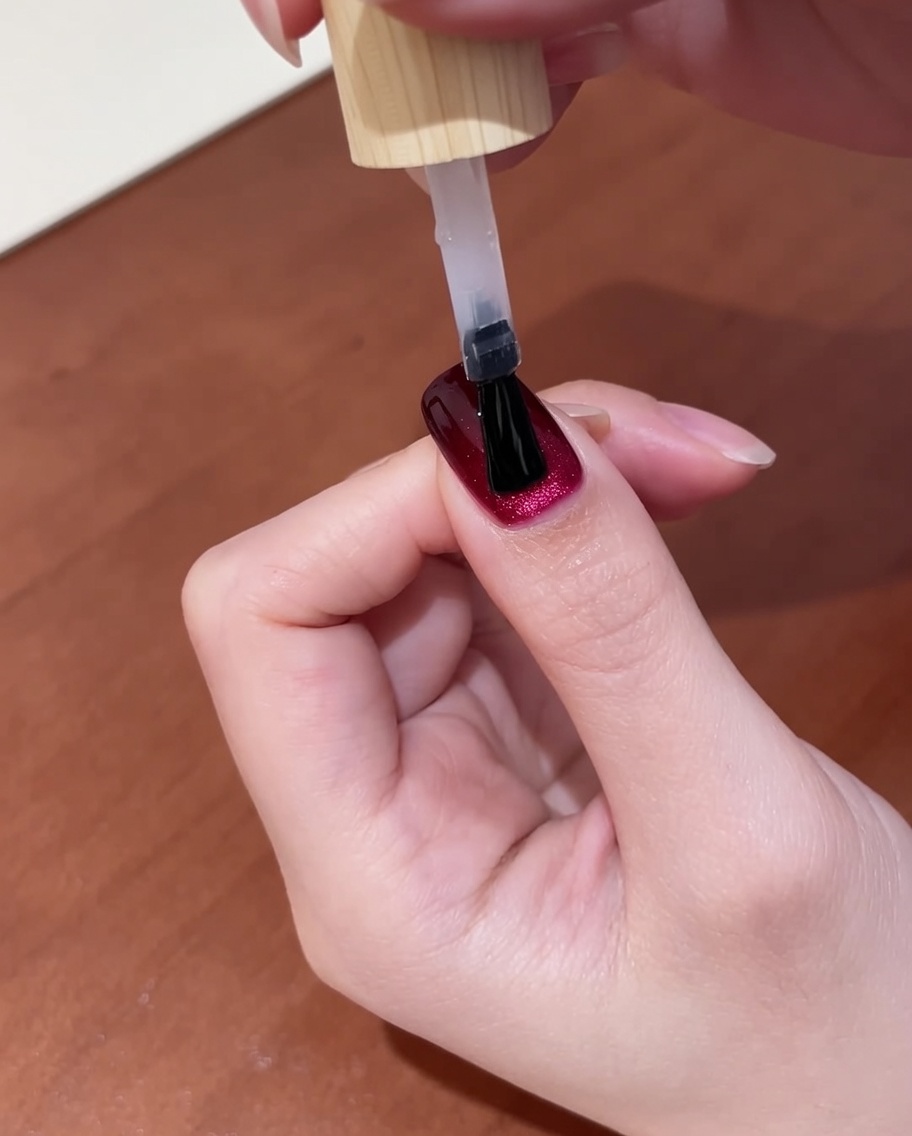 Nahaufnahme einer Hand mit frisch aufgetragenem Top-Coat auf dunkelroten Cat-Eye-Nägeln, bereit für den letzten Aushärtungsschritt in der Lampe.