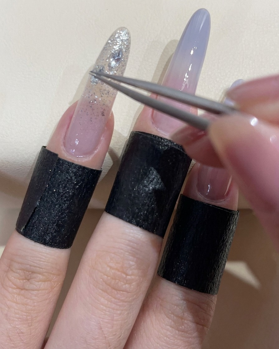 Eine Pinzette platziert einen glitzernden, klaren Zirkonstein auf den Nagel mit Klebegel.