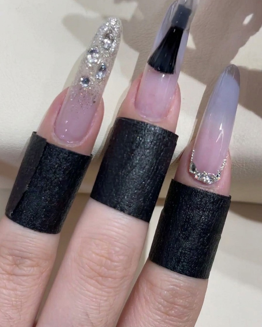 Ein schwarzer Pinsel trägt klaren Überlack auf einen Ombré-Nagel auf, während benachbarte Finger bereits mit Glitzer und Strass verziert sind.