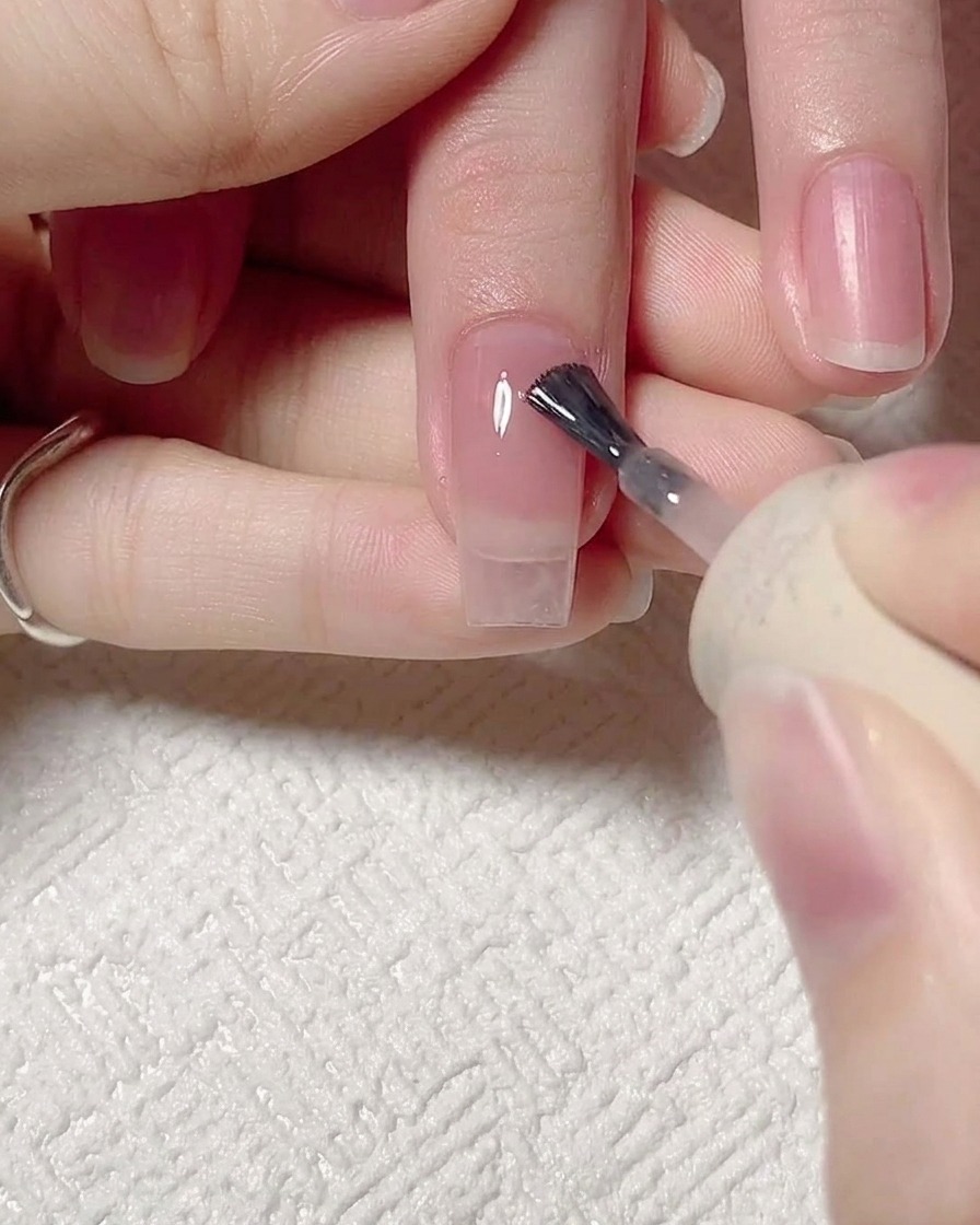 Nahaufnahme eines Pinsels, der ein transparentes rosa Jelly-Gel auf einen vorbereiteten Naturnagel aufträgt.